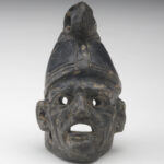 A small, black ceramic lantern in the shape of a man’s head wearing a cone-shaped hat. The face has deep wrinkles. There are holes in the eyes and under the ears, and the mouth is wide open. Front view.
