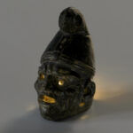 A small, black ceramic lantern in the shape of a man’s head wearing a cone-shaped hat. The face has deep wrinkles. There are holes in the eyes and under the ears, and the mouth is wide open. View of lamp lit with an LED light.