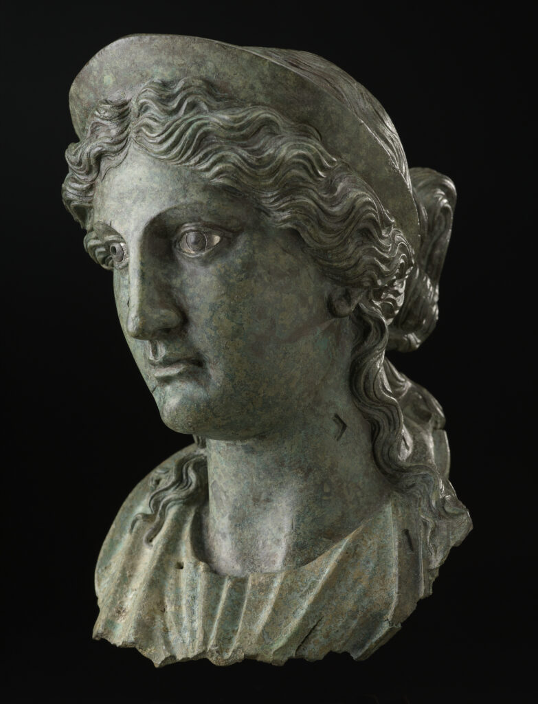 A bronze metal sculpture of a woman’s head and neck. She is wearing a crown on top of her head and has wavy hair worn in an updo.