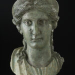 A bronze metal sculpture of a woman’s head and neck. She is wearing a crown on top of her head and has wavy hair worn in an updo. View of front.