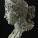 A bronze metal sculpture of a woman’s head and neck. She is wearing a crown on top of her head and has wavy hair worn in an updo. Side view.
