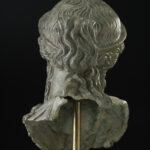A bronze metal sculpture of a woman’s head and neck. She is wearing a crown on top of her head and has wavy hair worn in an updo. View of back.
