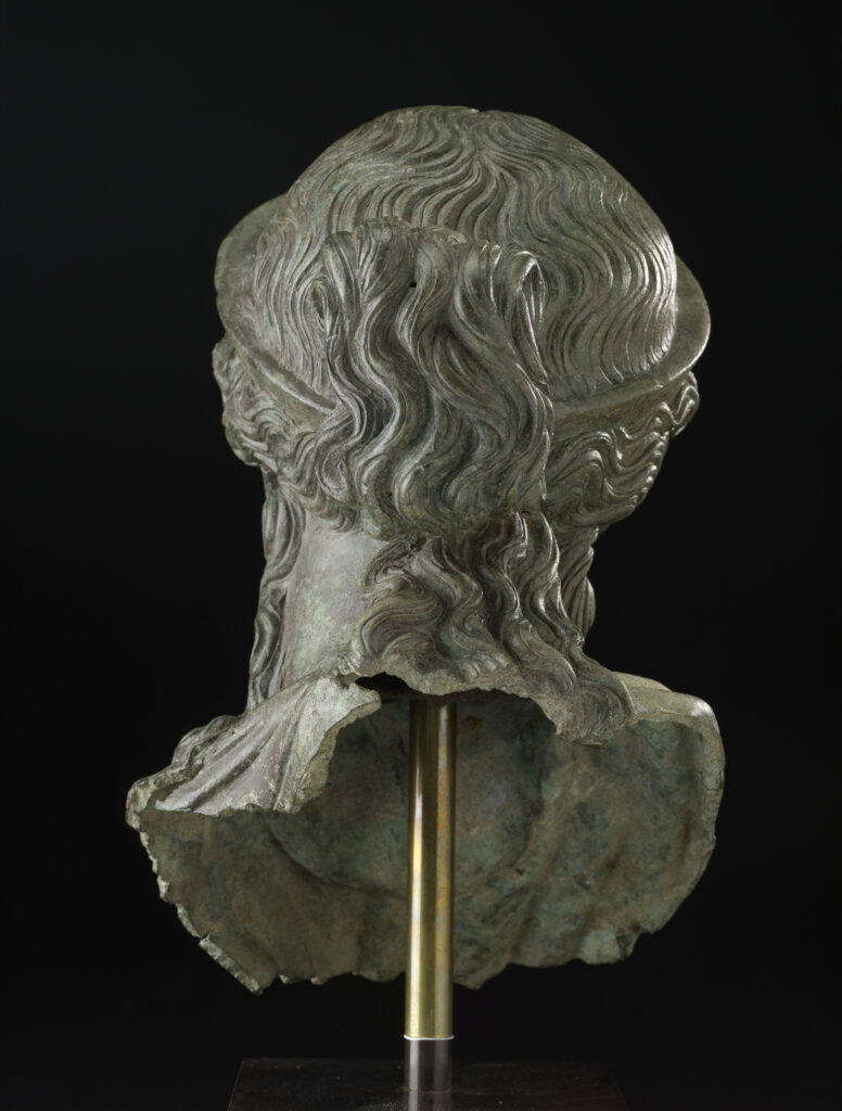 A bronze metal sculpture of a woman’s head and neck. She is wearing a crown on top of her head and has wavy hair worn in an updo.