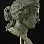 A bronze metal sculpture of a woman’s head and neck. She is wearing a crown on top of her head and has wavy hair worn in an updo. Side view.