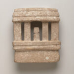 A stone sculpture of a temple with a human-like figure standing between the columns. View of reverse.