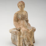 A clay sculpture of a seated woman dressed in a long, sleeveless gown.