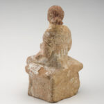 A clay sculpture of a seated woman dressed in a long, sleeveless gown. View of back.