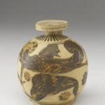 A beige-colored clay pot with a round base, a small neck, a handle, and flat mouth. It is decorated with black painted designs of flowers, a dolphin, and geometric shapes.