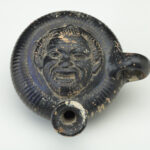 A black ceramic flask with a man’s face featured in the center. View from above.