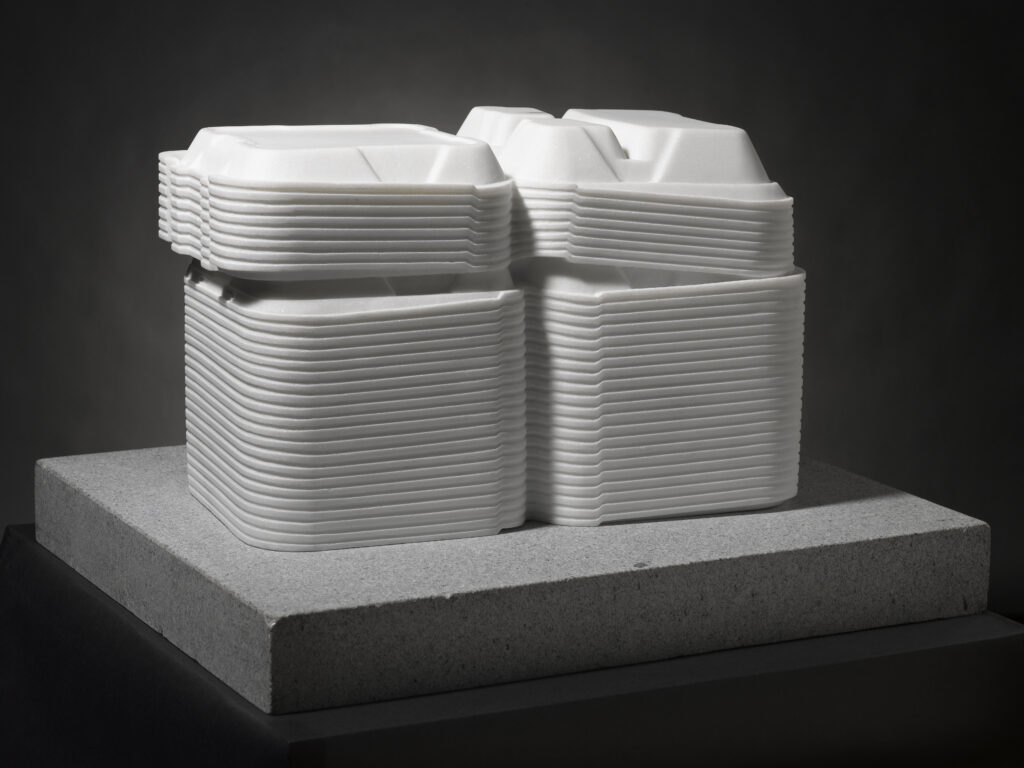 A white marble sculpture of open Styrofoam takeout boxes stacked on top of a gray, rectangle-shaped base.