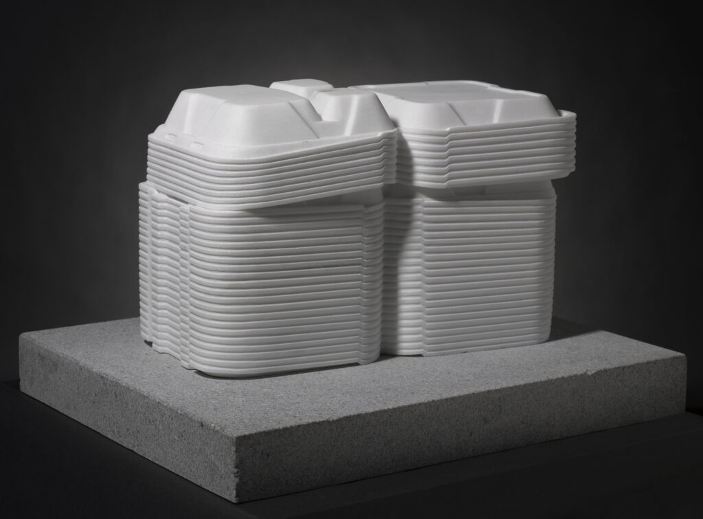 A white marble sculpture of open Styrofoam takeout boxes stacked on top of a gray, rectangle-shaped base.