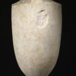 A cylinder-shaped marble vessel with a tapered top and a tapered base. The center of the vessel is engraved with the design of a seated man and a standing woman shaking hands. View of back.