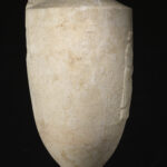 A cylinder-shaped marble vessel with a tapered top and a tapered base. The center of the vessel is engraved with the design of a seated man and a standing woman shaking hands. Side view.