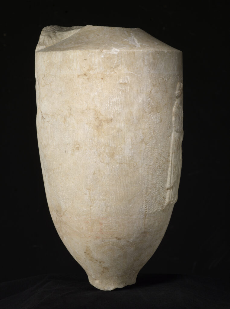 A cylinder-shaped marble vessel with a tapered top and a tapered base. The center of the vessel is engraved with the design of a seated man and a standing woman shaking hands.