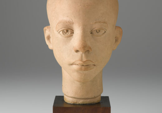 A terracotta sculpture of a boy’s head and neck on top of a brown, wooden, cube-shaped base. View of the front.