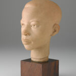 A terracotta sculpture of a boy’s head and neck on top of a brown, wooden, cube-shaped base. View of the face and part of the side.