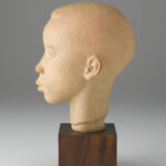 A terracotta sculpture of a boy’s head and neck on top of a brown, wooden, cube-shaped base. Side view.