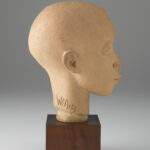A terracotta sculpture of a boy’s head and neck on top of a brown, wooden, cube-shaped base. Side view.