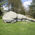 A large gray sculpture of a humanlike figure lying on its side outdoors