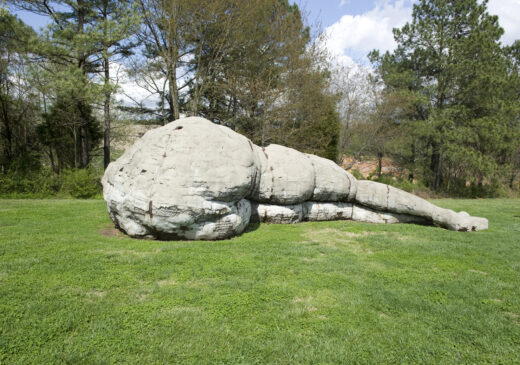 A large gray structure resembling the lower torso, legs, and feet of a human figure. The figure is lying on one side, facing away, on a grassy lawn with tall pine trees in the background.