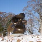 A bronze abstract sculpture in a field, with trees and a blue sky in the background. The ground is covered in snow and the trees are missing most of their leaves.