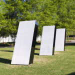 An outdoor image of three identical silver structures. They are positioned at an angle, in a single row, in front of tall trees on a grassy lawn.