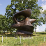 A bronze abstract sculpture in a field, with trees and a blue sky in the background.