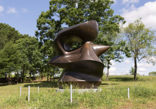 A bronze abstract sculpture in a field, with trees and a blue sky in the background.