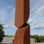 An image of a rust-colored abstract sculpture in a field, in front of a building and a blue sky. The sculpture is roughly rectangular and is made up of two divided sections of a single form. The larger form appears to balance on top of the smaller form.