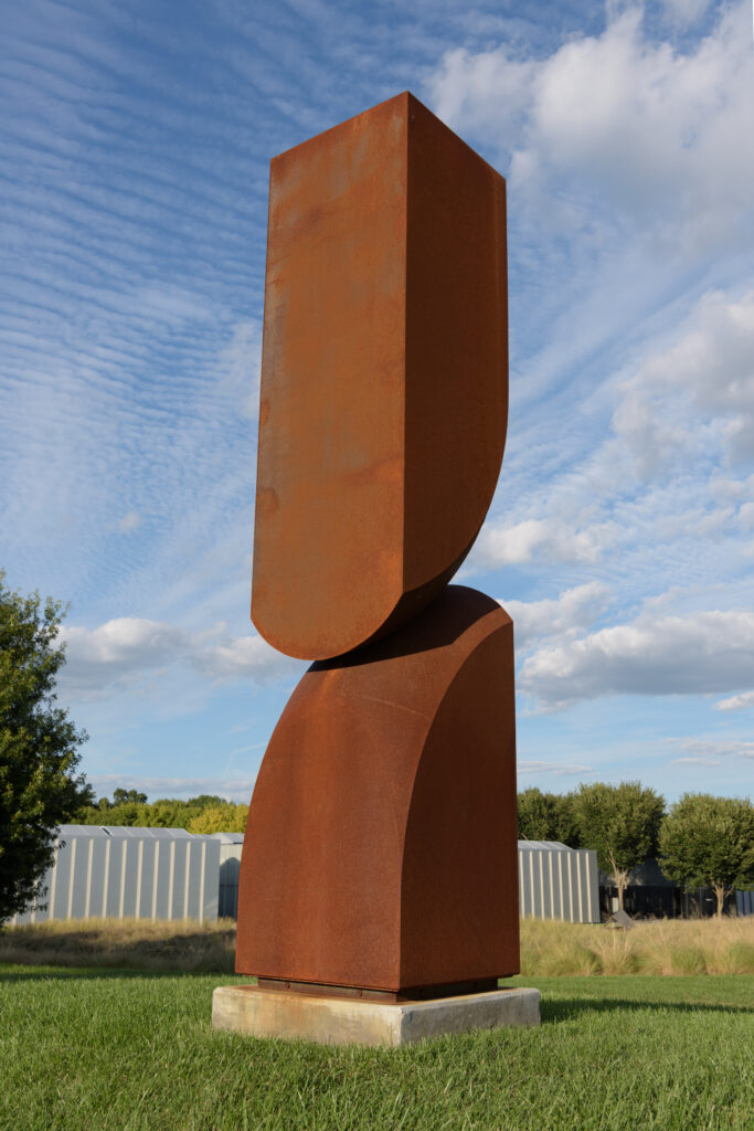 An image of a rust-colored abstract sculpture in a field, in front of a building and a blue sky. The sculpture is roughly rectangular and is made up of two divided sections of a single form. The larger form appears to balance on top of the smaller form.