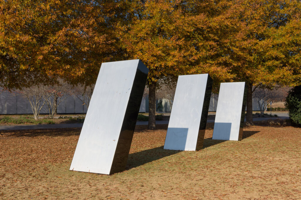 An outdoor image of three identical silver structures. They are positioned at an angle, in a single row, in front of tall trees on a grassy lawn.