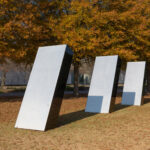 An outdoor image of three identical silver structures. They are positioned at an angle, in a single row, in front of tall trees on a grassy lawn. It is fall, the leaves on the trees are shades of orange and red, and leaves cover the ground around the sculptures.