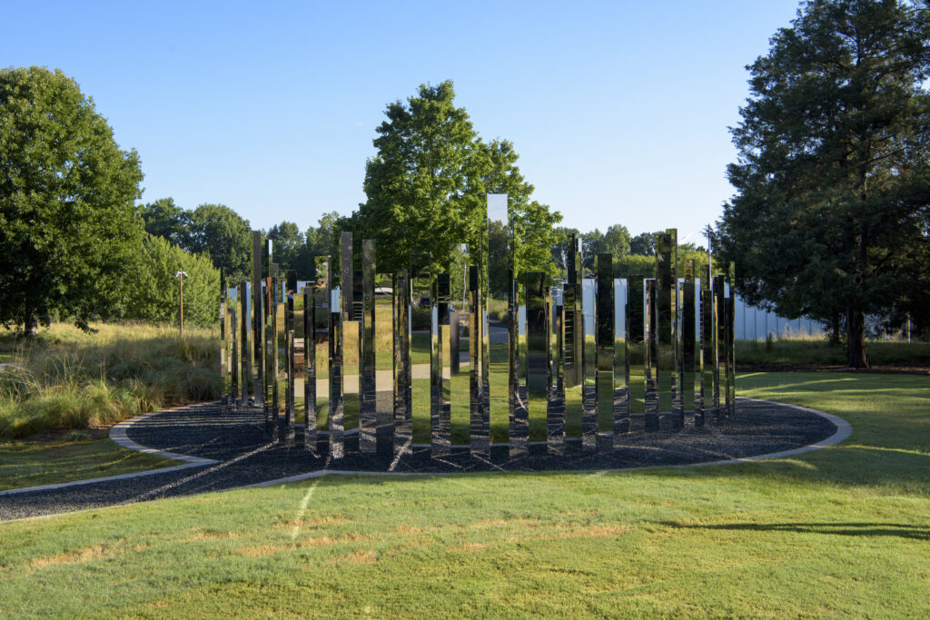 An outdoor sculpture of a circular maze made up of vertical mirrored panels. The panels stand upright inside a gravel circle surrounded by green grass. There are tall green trees and a clear blue sky in the background.