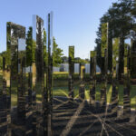 An outdoor sculpture of a circular maze made up of vertical mirrored panels. The panels stand upright inside a gravel circle surrounded by green grass. There are tall green trees and a clear blue sky in the background. View of the interior of the mirrored circle.