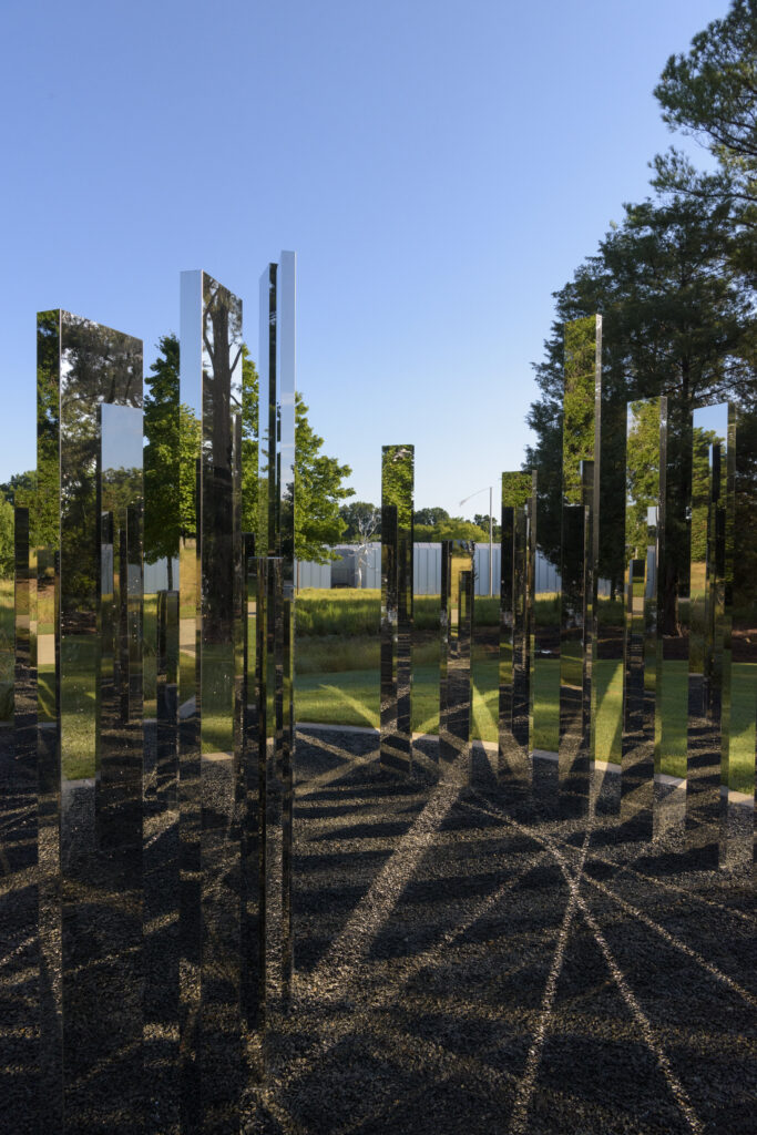 An outdoor sculpture of a circular maze made up of vertical mirrored panels. The panels stand upright inside a gravel circle surrounded by green grass. There are tall green trees and a clear blue sky in the background.