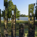 An outdoor sculpture of a circular maze made up of vertical mirrored panels. The panels stand upright inside a gravel circle surrounded by green grass. There are tall green trees and a clear blue sky in the background. Close up view of mirrored panel.