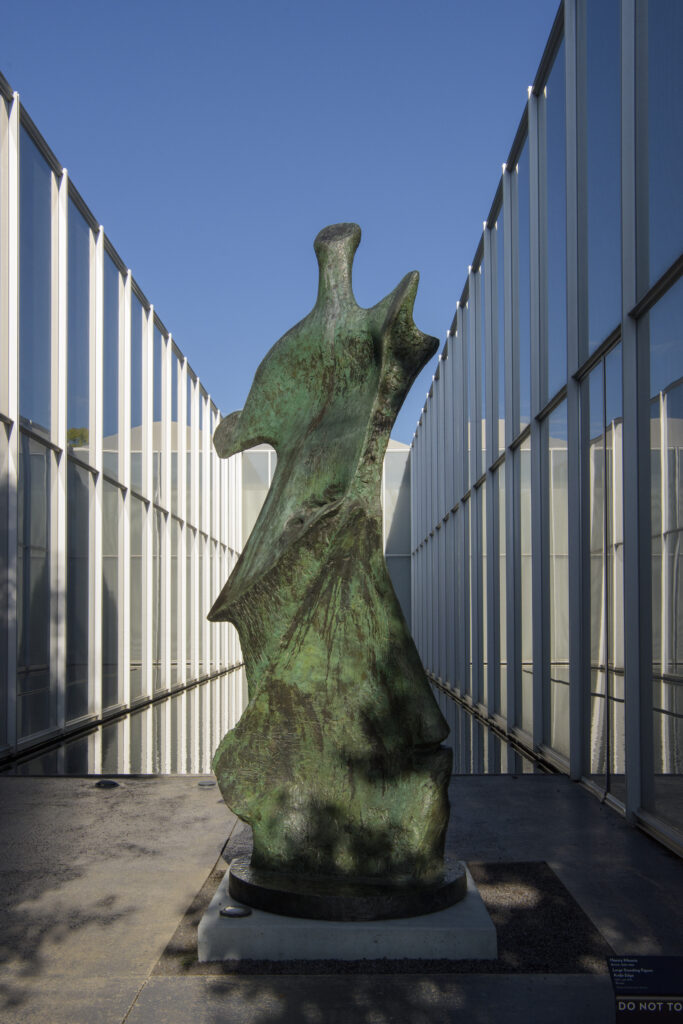 A tall, greenish-brown abstract sculpture with a bonelike form and jutting edges on each side. A glass building, a small section of blue sky, and the edge of a reflecting pool are in the background.
