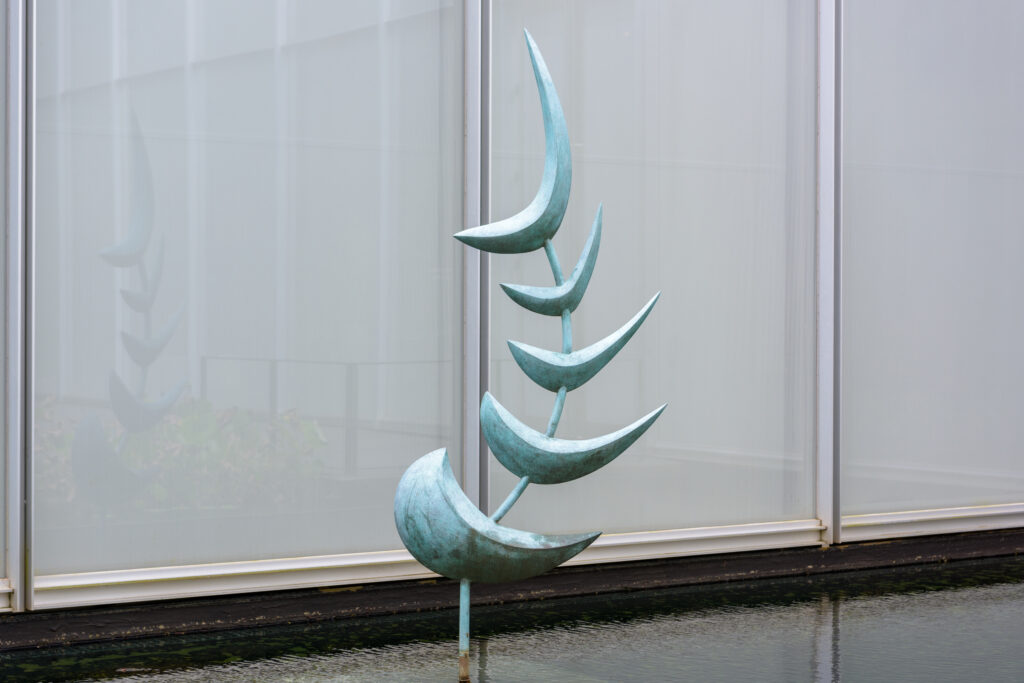 A metal abstract fishlike sculpture standing upright in a shallow pool of water. The sculpture is made up of five blue-green crescent shapes on a curved pole. There are windows in the background and aquatic plants in the foreground.
