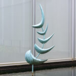 A metal abstract fishlike sculpture standing upright in a shallow pool of water. The sculpture is made up of five blue-green crescent shapes on a curved pole. There are windows in the background. View from the front.