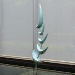 A metal abstract fishlike sculpture standing upright in a shallow pool of water. The sculpture is made up of five blue-green crescent shapes on a curved pole. There are windows in the background. View from the side.