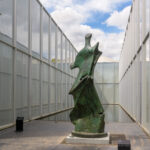 A tall, greenish-brown abstract sculpture with a bonelike form and jutting edges on each side. A glass building, a small section of blue sky, and the edge of a reflecting pool are in the background.