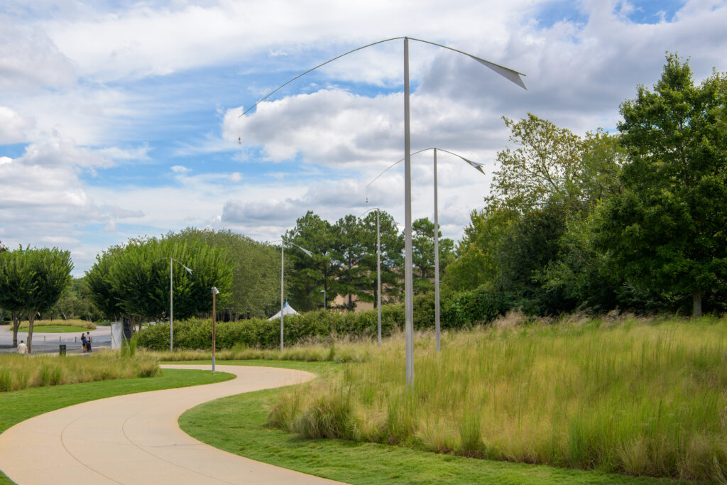 An outdoor image of a grassy lawn with five metal posts lining a curved sidewalk. Each post supports a curved beam with a winged form attached at the top on one side, and a small bell is suspended from the other side.