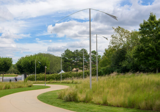 An outdoor image of a grassy lawn with five metal posts lining a curved sidewalk. Each post supports a curved beam with a winged form attached at the top on one side, and a small bell is suspended from the other side.