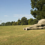 A large gray sculpture of a humanlike figure lying on its side outdoors