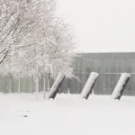 An outdoor image of three identical silver structures. They are positioned at an angle, in a single row, in front of a glass and metal building. Their is a tree to the left, and everything is covered in snow.