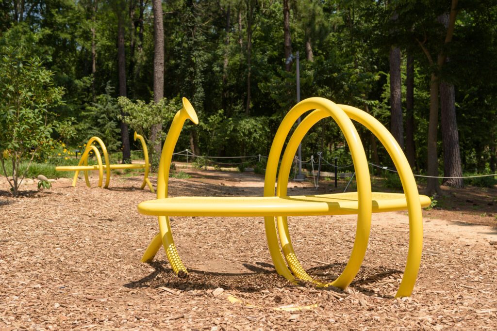 An image of two yellow benches on a mulch clearing in front of trees. The benches are encircled by tubes that support the bench. Each tube has a disk-shaped opening.