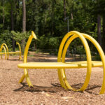 An image of two yellow benches on a mulch clearing in front of trees. The benches are encircled by tubes that support the bench. Each tube has a disk-shaped opening.