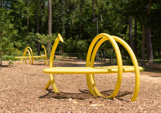 An image of two yellow benches on a mulch clearing in front of trees. The benches are encircled by tubes that support the bench. Each tube has a disk-shaped opening.