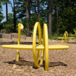An image of two yellow benches on a mulch clearing in front of trees. The benches are encircled by tubes that support the bench. Each tube has a disk-shaped opening.