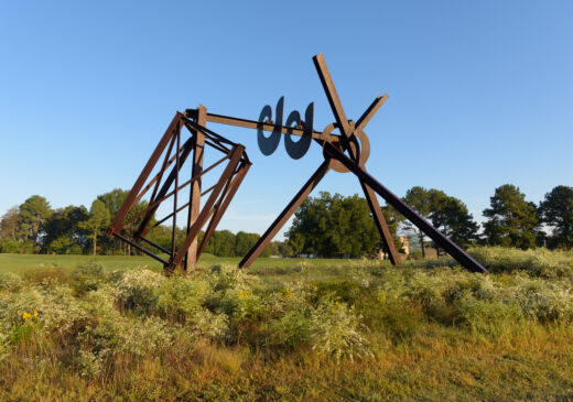 A large, brown metal structure in a grassy field. It has five angled beams that connect with circular forms and a three-dimensional rectangle shape that appears to balance on a single point. ​​